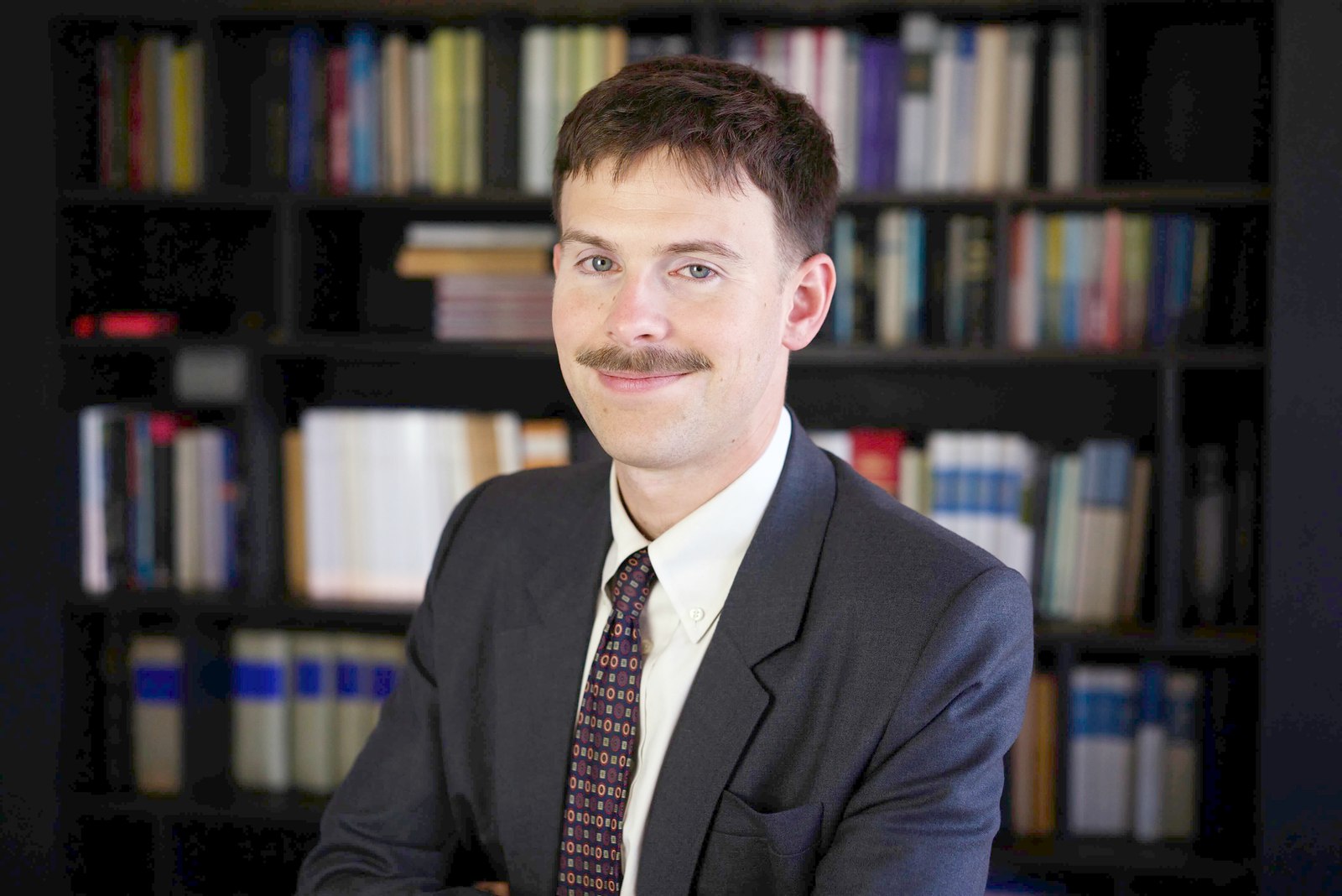 Corporate headshot of a smiling man with a moustache in front of a bookshelf