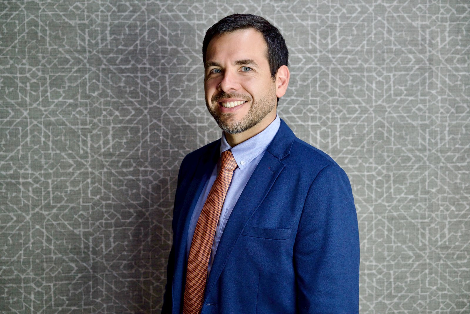 Corporate headshot of a smiling man with a beard and an orange tie