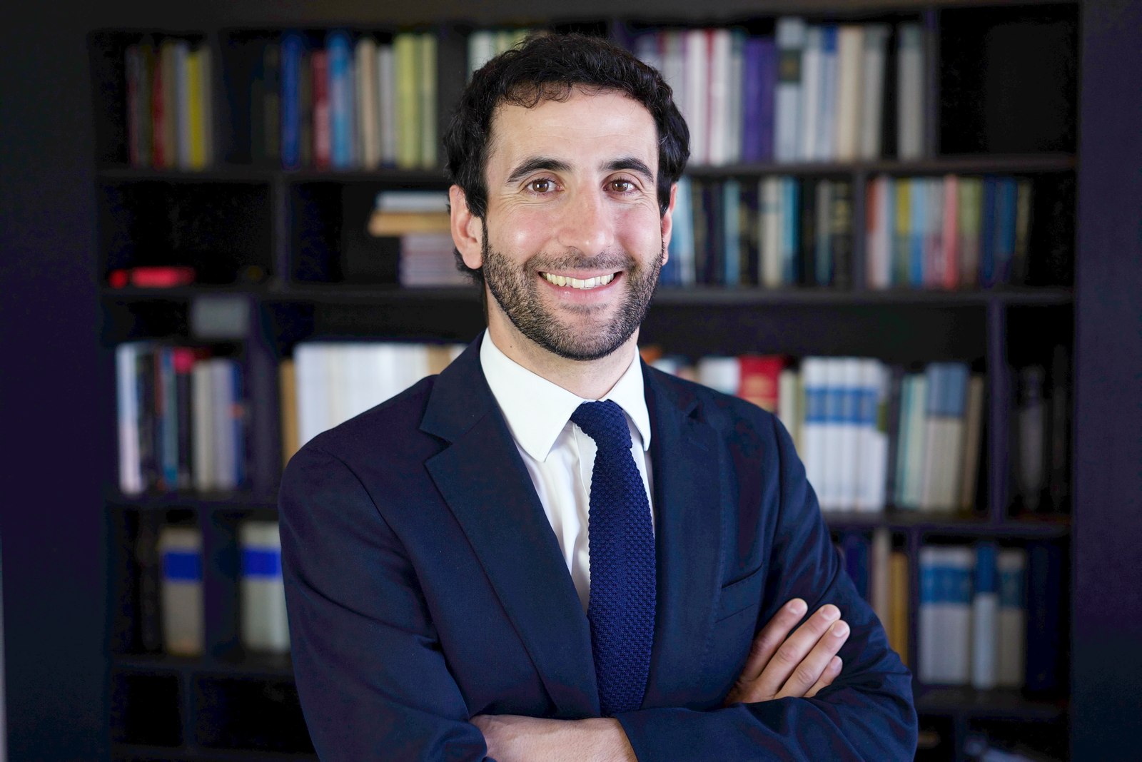 Corporate headshot of a smiling bearded man in front of a bookshelf, arms crossed