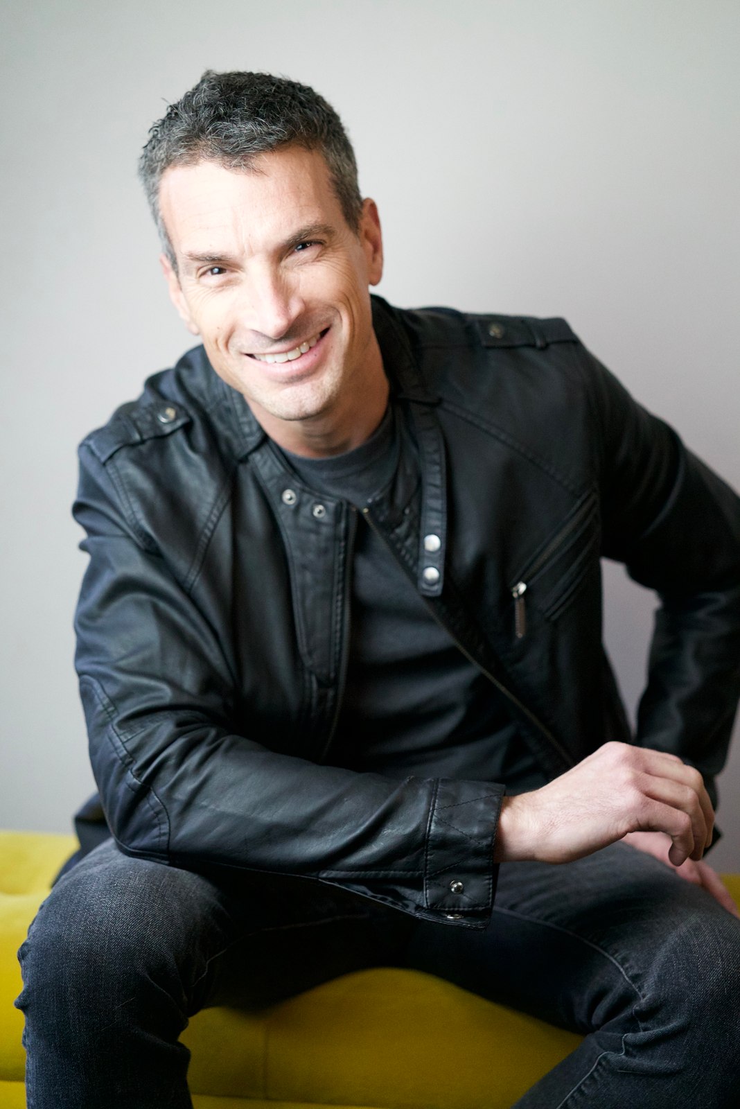 Portrait of a smiling man in a black leather jacket seated on a yellow bench